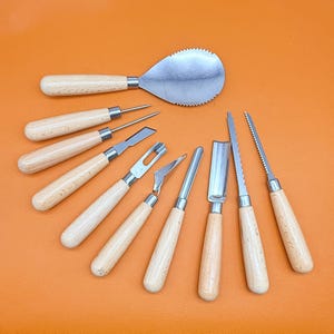 Könnte beinhalten: Ein Set aus acht Schnitz- und Modellierwerkzeugen mit hellen Holzgriffen und silberfarbenen Metallklingen. Die Sammlung umfasst einen gezackten Löffel, zwei spitze Ahlen, einen Flachmeißel, ein U-förmiges Hohleisen und verschiedene Sägeklingen.