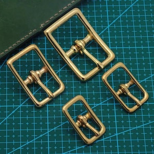 Könnte beinhalten: Vier rechteckige Messing-Gürtelschnallen mit Rollensteg. Die Schnallen sind in verschiedenen Größen und auf einer grün-blauen Rasteroberfläche angeordnet. Das Metall hat eine glänzende, goldene Oberfläche.