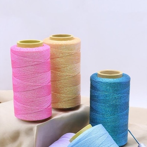 Peut inclure: Plusieurs bobines de fil colorées, rose, doré et bleu. Le fil est enroulé autour d'un noyau jaune, formant une forme cylindrique. Les bobines sont disposées sur une surface satinée.