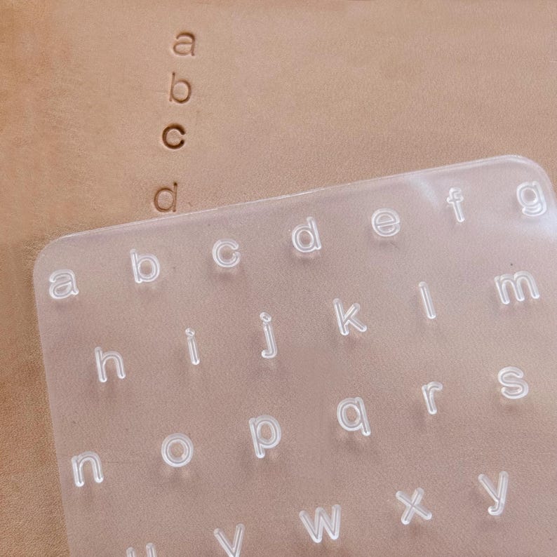 May include: A clear plastic alphabet stamp set with lowercase letters. The letters are arranged in rows and columns. The image also shows the letters "a", "b", "c", and "d" stamped into a piece of tan leather.