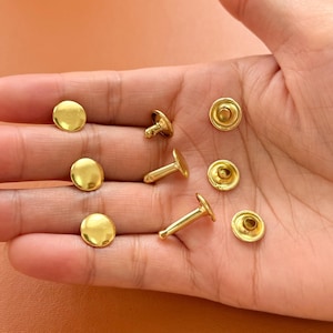 Puede incluir: Primer plano de remaches y cierres dorados. Los remaches tienen una cabeza plana y redonda y un poste para asegurar los materiales. Los cierres tienen una cabeza circular con un centro empotrado. Los artículos se muestran en una mano.