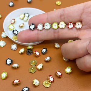 May include: A selection of small, colourful penguin-shaped stud earrings. The earrings feature a variety of colours, including black, white, blue, green, and red, with gold-coloured metal accents. Some earrings are in a small white dish.