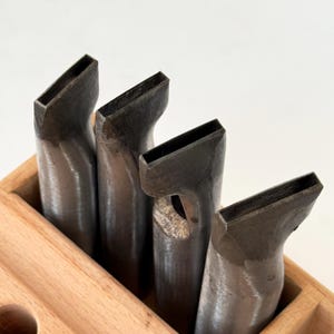 Puede incluir: Cuatro herramientas metálicas para trabajar el cuero con aberturas rectangulares, dispuestas en un soporte de madera. Las herramientas tienen un acabado metálico oscuro y están anguladas. El soporte de madera es marrón claro.