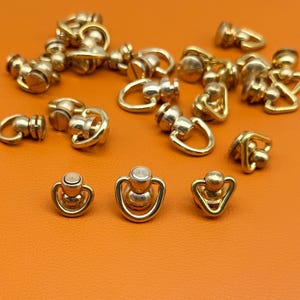 Puede incluir: Una colección de pequeños remaches en forma de anilla en D de color dorado metálico con mecanismo de tornillo. Cada pieza presenta una base redondeada, un poste central y un lazo triangular unido a una cabeza circular para cuero o tela.