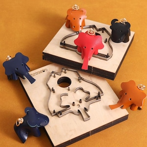 Peut inclure: Des porte-clés en cuir en forme d'éléphants et de taureaux, de couleurs variées (orange, rouge, bleu, noir), sont présentés avec des gabarits de découpe en bois. Les porte-clés ont des attaches dorées et sont disposés sur un fond jaune-orangé.