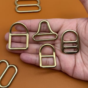 May include: A collection of gold-colored metal hardware pieces, including D-rings, rectangular rings, and a unique trapezoid-shaped piece. The hardware is displayed against a warm, orange-brown background, suggesting a craft or design context.