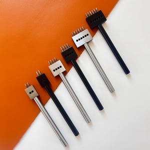 Puede incluir: Una colección de herramientas de marroquinería, incluyendo punzones de costura de varios tamaños. Las herramientas tienen mangos de metal plateado y cabezas negras o plateadas con múltiples púas. El fondo es una mezcla de naranja y blanco.