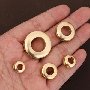 Peut inclure: Plusieurs oeillets métalliques dorés de différentes tailles. Les oeillets circulaires ont un trou central et sont présentés sur une main. Ces pièces de quincaillerie sont probablement utilisées pour des projets d'artisanat, de couture ou de maroquinerie.