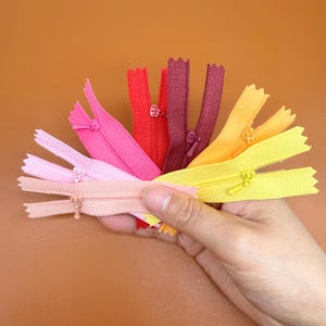 Puede incluir: Una mano sostiene un abanico de cremalleras de colores en tonos rosa, rojo, amarillo y morado. Las cremalleras están abiertas, mostrando sus dientes y tiradores. El fondo es de color marrón liso.