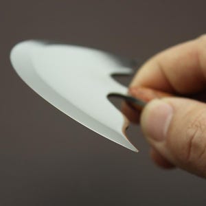 Op de afbeelding: Close-up van een klein, zilverkleurig, bladvormig gereedschap met een scherpe rand. Het gereedschap wordt door een hand vastgehouden en de achtergrond is effen en neutraal van kleur. Het gereedschap lijkt van metaal te zijn.
