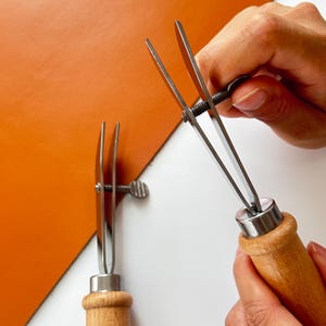 May include: Two leathercraft tools with wooden handles and adjustable metal blades are shown. One tool is being adjusted by a hand. A piece of orange leather is in the background. The tools are used for leatherworking.