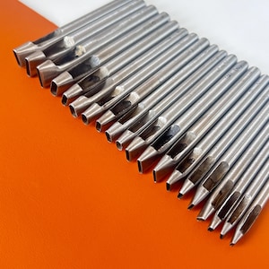 Könnte beinhalten: Eine Sammlung von silbernen Metall-Lochstempeln für Lederarbeiten. Jeder Stempel hat ein rechteckiges Schneidende und eine hohle Mitte. Die Werkzeuge sind diagonal vor einem orangefarbenen Hintergrund angeordnet.