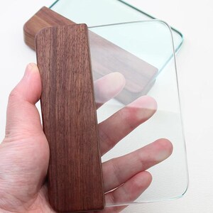 May include: A clear glass cutting board with a dark brown wooden handle. The handle is rectangular and appears to be made of walnut. The cutting board is rectangular with rounded corners. The image is taken from a close-up angle.
