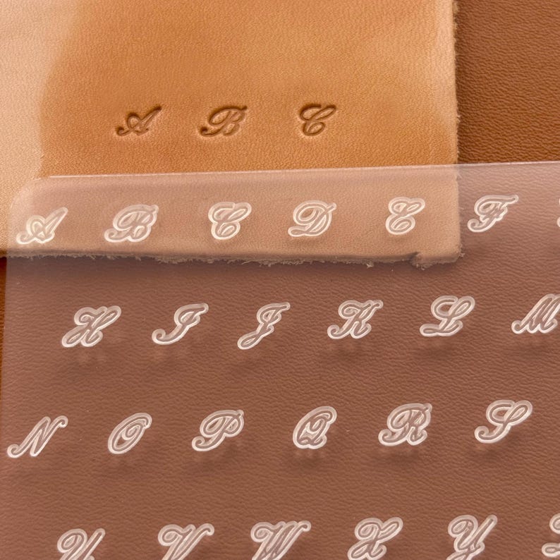 May include: Close-up of leather stamping tools with the letters A through Y. The letters are in a cursive font and are displayed on a clear plastic sheet. Stamped letters A, B, and C are visible on the leather.