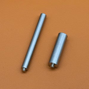 May include: Two cylindrical metal components with a smooth, polished silver-toned finish. One component is a long, slender rod, while the other is a shorter, thicker cylinder. Both pieces feature a small, threaded screw end for attachment.
