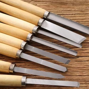 May include: A set of wood carving tools with wooden handles and silver-colored blades. The blades have various shapes, including flat, angled, and pointed tips. The tools are arranged on a wooden surface.