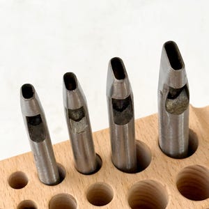May include: Four metal hole punch tools of varying sizes are arranged in a wooden holder. The tools have a cylindrical body with a flat, angled cutting edge. The wooden holder has several round holes for the tools and other items.