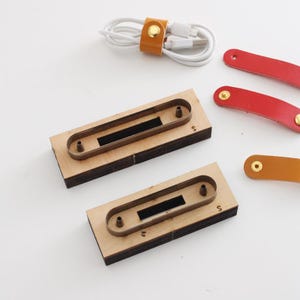 Op de afbeelding: Een bovenaanzicht toont diverse lederen ambachtelijke gereedschappen en accessoires. Er zijn twee houten mallen met metalen frames, verschillende leren riemen in rood en oranje, en een kabelorganizer. De kabelorganizer is gemaakt van oranje leer met een gouden drukknop.