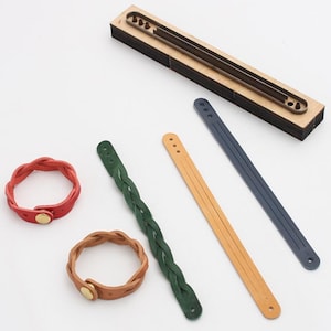 Può includere: Una vista dall'alto mostra componenti per braccialetti in pelle e braccialetti finiti. I componenti includono una fustella in legno e strisce di pelle blu, beige e verde. Due braccialetti finiti sono rossi e marroni, con bottoni automatici dorati.