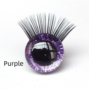 May include: A purple, glitter-encrusted glass eye with black eyelashes. The word "Purple" is printed in black text below the eye. The eye reflects the surrounding environment.