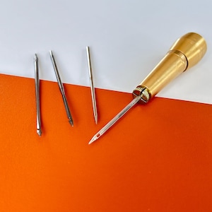 May include: A brass handle awl with a silver needle inserted, plus three spare silver needles. The awl and needles are displayed on a white and orange surface, suitable for leatherwork and sewing.