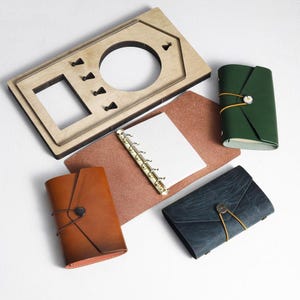 May include: An overhead shot displays a collection of leather-bound notebooks in various colors: brown, green, and navy. A wooden frame with cutouts and a ring-bound notebook with grid paper are also present. The notebooks are closed with a button and string.