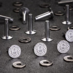 Könnte beinhalten: Nahaufnahme verschiedener Metallnieten und Unterlegscheiben. Einige Nieten haben einen dekorativen kreisförmigen Kopf mit einem Sonnenstrahl-Design und einer zentralen Vogelfigur. Die Nieten sind silberfarben und die Unterlegscheiben sind aus dunklerem Metall.