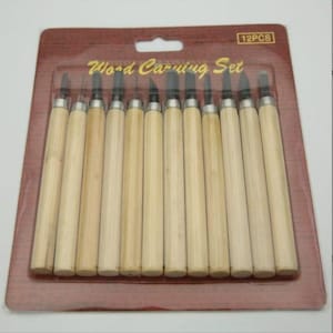 Pode incluir: Um conjunto de 12 ferramentas para entalhar madeira com cabos de madeira e lâminas de metal, embaladas em uma embalagem blister transparente. A embalagem está em uma base de papelão marrom com a inscrição "Wood Carving Set".