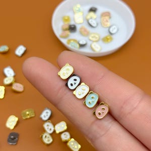 May include: A collection of small, colorful panda-shaped charms. The charms are in various colors, including black and white, light blue, and pink, with gold-colored borders. Some charms are on a finger, while others are in a white dish.
