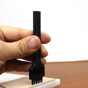 Può includere: Uno strumento di scalpello per cuciture in pelle nera con più rebbi. Lo strumento è tenuto sopra un pezzo di pelle marrone chiaro, pronto a praticare fori per cucire. Lo sfondo è una superficie marrone chiaro.