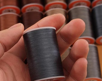 May include: A spool of dark gray thread is held in a hand, with a blurred background of other thread spools in various shades of brown and gray. The thread is tightly wound on a white and orange spool.