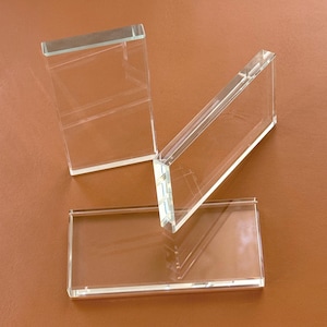 May include: Three clear, rectangular glass blocks of varying sizes are arranged on a brown surface. The blocks are transparent, allowing light to pass through, and have a clean, modern aesthetic. The edges are smooth and well-defined.