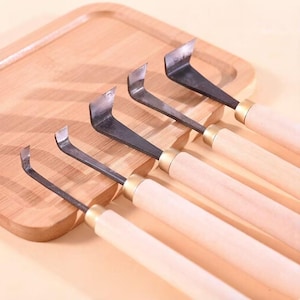 Puede incluir: Un juego de cinco herramientas para tallar madera con cuchillas curvas de metal negro y mangos de madera de color claro. Las herramientas están dispuestas sobre una bandeja de madera marrón claro.