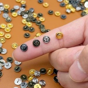 Peut inclure: Un assortiment de petits boutons ronds aux finitions métalliques variées, or, argent et noir. Chaque bouton présente un motif d'ancre détaillé. Les boutons sont dispersés sur une surface marron.