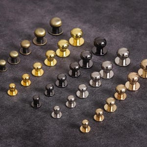 May include: An assortment of metal rivets in various sizes and finishes, including gold, brass, silver, and black. The rivets have a rounded head and a flat base, suitable for leathercraft or garment construction.