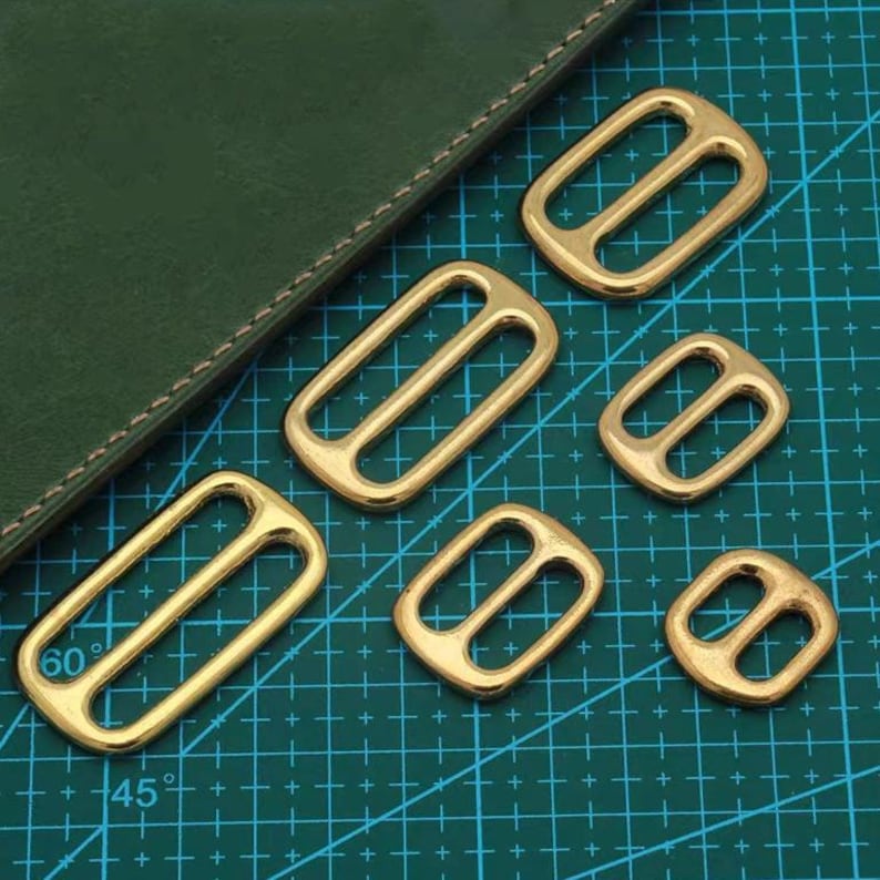 Op de afbeelding: Een verzameling goudkleurige metalen schuifgespen in diverse maten, geplaatst op een groene rasterachtergrond. De gespen hebben een rechthoekige of afgeronde vorm met een centrale staaf.