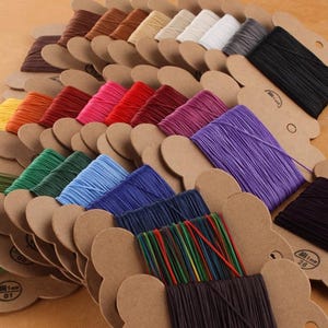 May include: A selection of colourful, 1mm diameter waxed cotton cords, neatly arranged on brown cardboard spools. The cords come in a variety of colours, including shades of brown, beige, white, grey, black, orange, red, pink, purple, green, and blue.