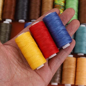 Puede incluir: Una mano que sostiene tres carretes de hilo en amarillo, rojo y azul. Los carretes están bien enrollados con una textura visible. Otros carretes de hilo en varios colores están en el fondo, incluyendo marrón, verde y turquesa.