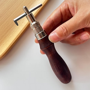 May include: A leathercraft edge beveler tool featuring a dark brown wooden handle, a silver metal body, and a sharp, angled blade. This tool is designed for shaping and finishing leather edges. The handle is ergonomically shaped for a comfortable grip.