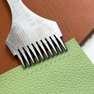 May include: A silver-colored metal leather stitching chisel with nine sharp prongs. The tool is positioned over a piece of light green textured leather, showing a row of nine small, evenly spaced slits punched into the material surface.