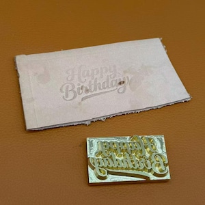 May include: A rectangular brass metal stamp featuring the embossed text Happy Birthday in a cursive script, positioned below a light-coloured rectangular leather tag that displays the same Happy Birthday text imprinted into its surface.