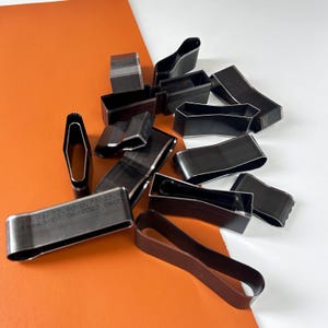 Puede incluir: Una colección de diversas herramientas de artesanía en cuero de metal negro, que incluyen formas rectangulares, curvas y ovaladas. Algunas herramientas tienen texto estampado. Las herramientas están dispuestas sobre un fondo naranja y blanco.