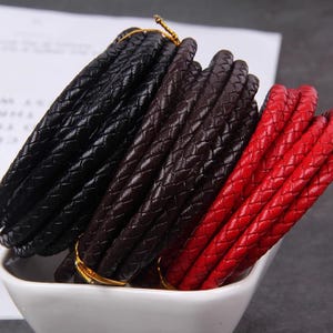 May include: Braided leather cords in black, brown, and red are bundled together. The cords are tightly woven, and the ends are secured with gold-colored ties. The cords are displayed in a white bowl, suggesting a craft or jewelry supply.