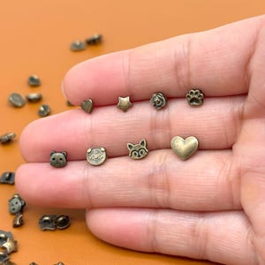 Könnte beinhalten: Eine Sammlung kleiner, antik-messingfarbener Metallverzierungen in verschiedenen Formen, darunter Herzen, Sterne, Blumen, Pfotenabdrücke, ein Bär, ein Schwein und eine Katze. Diese dekorativen Stücke sind ideal zum Basteln und für Detailarbeiten.