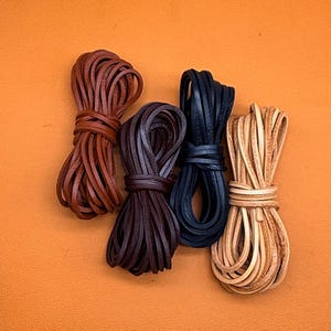 May include: Four bundles of flat leather cord arranged in a row. The colours from left to right are medium brown, dark brown, black, and natural tan. Each bundle is neatly coiled and secured with a matching leather strip wrapped around the centre.