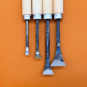May include: A set of four wood carving chisels with light-colored wooden handles and silver-toned metal ferrules. The steel blades vary in width and shape, ranging from a narrow straight edge to a wide, flared cutting edge with a metallic finish.