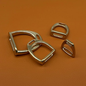 Puede incluir: Cuatro estribos plateados de diferentes tamaños están dispuestos sobre una superficie naranja. Los estribos tienen forma de D con una abertura rectangular en la parte inferior. El metal tiene un acabado pulido que refleja la luz.
