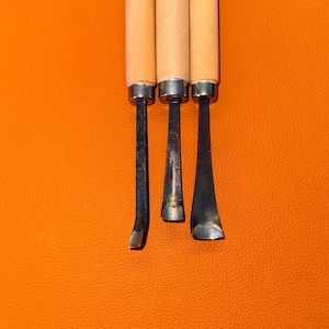 May include: Three wood carving tools with light-colored wooden handles and silver-toned metal ferrules. Each tool features a distinct steel blade shape, including a narrow straight chisel, a curved gouge, and a wider angled gouge for carving.