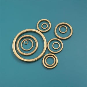 Op de afbeelding: Een verzameling goudkleurige metalen ringen van verschillende maten, geplaatst op een turquoise achtergrond. De ringen zijn cirkelvormig, met een gladde, gepolijste afwerking. De opstelling creëert een visueel aantrekkelijk patroon, geschikt voor knutselprojecten of decoratieve doeleinden.