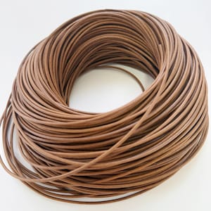May include: A tightly wound coil of brown leather cord. The cord has a smooth texture and a consistent thickness, with a warm, earthy hue. The coil is set against a white backdrop, accentuating the texture and colour of the leather.
